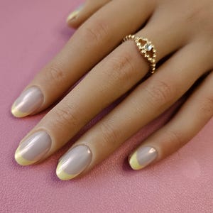Peut inclure: Une bague en or ornée d'une petite pierre ronde et transparente sertie sur un anneau perlé. La bague est portée sur un doigt aux ongles ovales, vernis d'une base brillante et claire avec des pointes distinctes en jaune pâle.