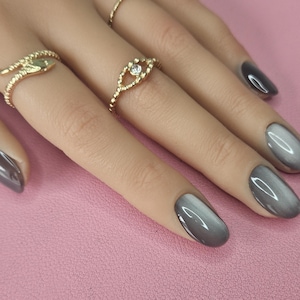 Könnte beinhalten: Nahaufnahme einer Hand mit grauen, oval geformten Nägeln und goldenen Ringen. Ein Ring hat ein Schlangendesign, ein anderer eine Augenform mit einem kleinen Edelstein. Der Hintergrund ist eine rosa Oberfläche.