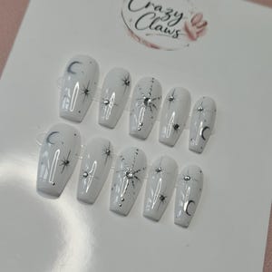Peut inclure: Un ensemble de dix faux ongles blancs à la finition brillante, ornés de motifs célestes argentés incluant des lunes, des étoiles et de petits strass. La carte de présentation en haut affiche le texte Crazy Claws avec une icône de main.