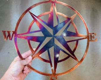 May include: A circular metal compass rose wall art piece featuring a star-shaped center with four cardinal direction points labeled N, S, E, and W. The metal surface displays a heat-treated finish with gradient tones of copper, blue, and purple.