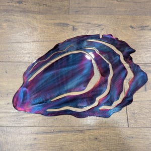 May include: A metal wall art piece shaped like an oyster shell with a swirling, abstract design in shades of blue, purple, and pink.