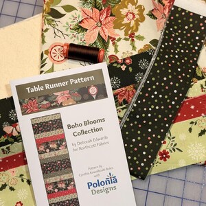 May include: A collection of fabric pieces and a sewing pattern for a table runner. The fabrics feature floral and geometric patterns in shades of green, red, and cream. The pattern is titled "Boho Blooms Collection" by Deborah Edwards for Northcott Fabrics.