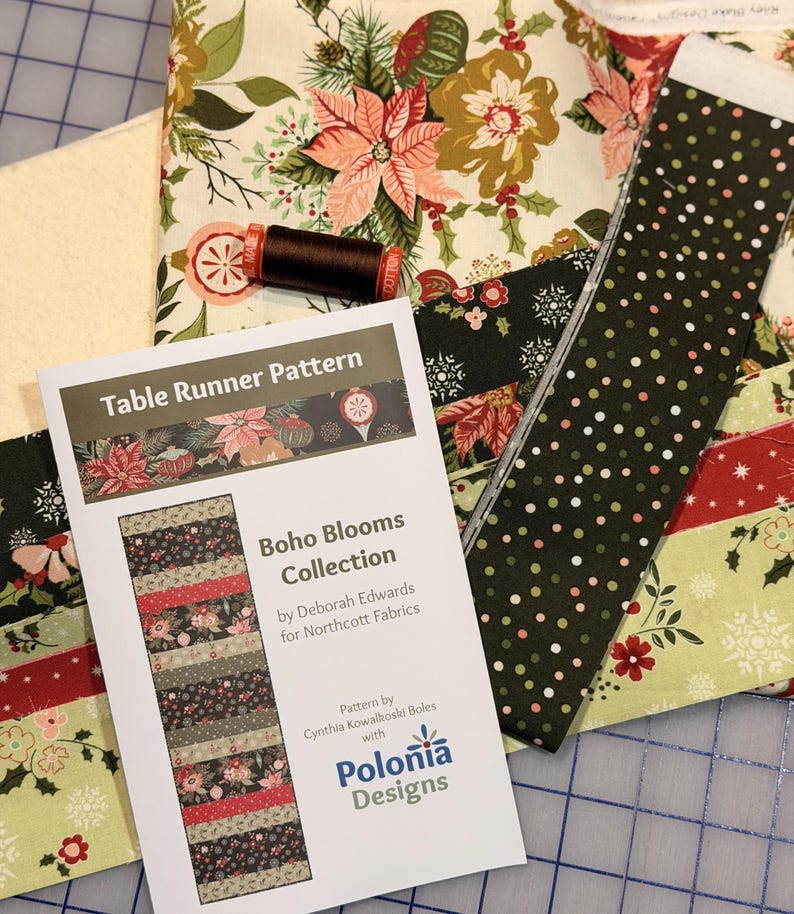 May include: A collection of fabric pieces and a sewing pattern for a table runner. The fabrics feature floral and geometric patterns in shades of green, red, and cream. The pattern is titled "Boho Blooms Collection" by Deborah Edwards for Northcott Fabrics.