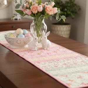 May include: A decorative Easter table setting featuring a floral table runner with pink and green patterns. Two white ceramic bunny figurines sit on the runner, alongside a bowl of pastel-colored Easter eggs and a vase of pink flowers.