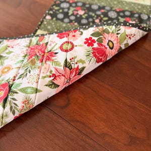 May include: A floral table runner with a variety of patterns and colors. The runner features a white background with red and pink flowers, green leaves, and a black border. The reverse side has a floral pattern with red poinsettias and green holly.
