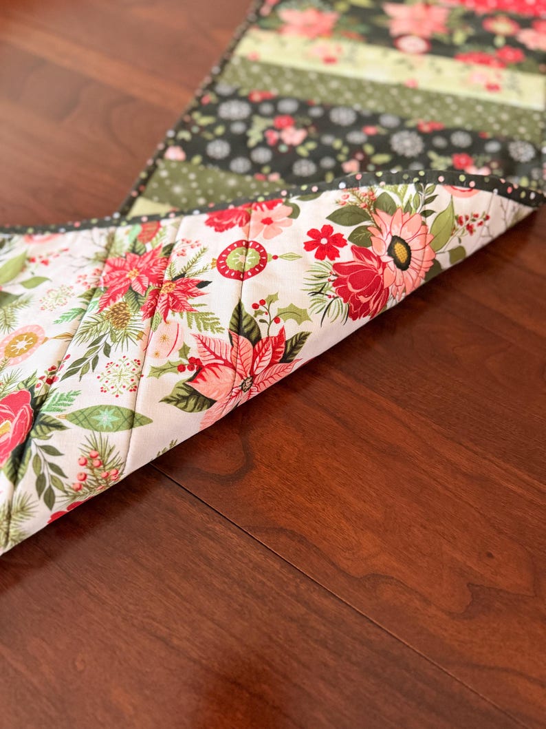 May include: A floral table runner with a variety of patterns and colors. The runner features a white background with red and pink flowers, green leaves, and a black border. The reverse side has a floral pattern with red poinsettias and green holly.