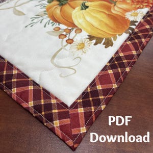 May include: A quilted table runner with a fall theme. The runner features a cream-colored center with a pumpkin and floral design. The border is a plaid pattern in shades of burgundy, gold, and brown. The text "PDF Download" is visible in the corner.