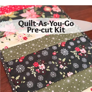 May include: A pre-cut quilt kit with a variety of patterned fabrics. The kit includes fabrics with floral, snowflake, and dotted designs in colors like red, black, green, and cream. The text "Quilt-As-You-Go Pre-cut Kit" is displayed.