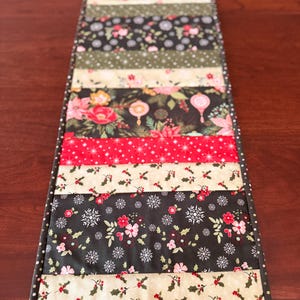 May include: A rectangular table runner with a patchwork design. The runner features alternating strips of fabric with floral, snowflake, and holly patterns in red, black, and cream colors. The edges are finished with a dotted border.