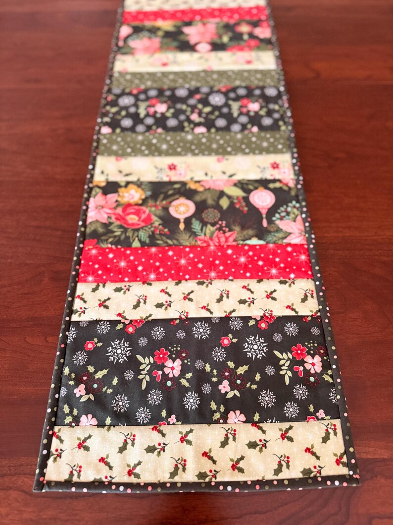 May include: A rectangular table runner with a patchwork design. The runner features alternating strips of fabric with floral, snowflake, and holly patterns in red, black, and cream colors. The edges are finished with a dotted border.