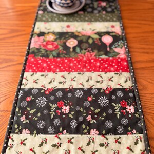 May include: A quilted table runner with a festive Christmas theme. The runner features alternating strips of fabric with floral, snowflake, and holly patterns in red, green, and cream colors. The edges are finished with a dark green polka dot border.