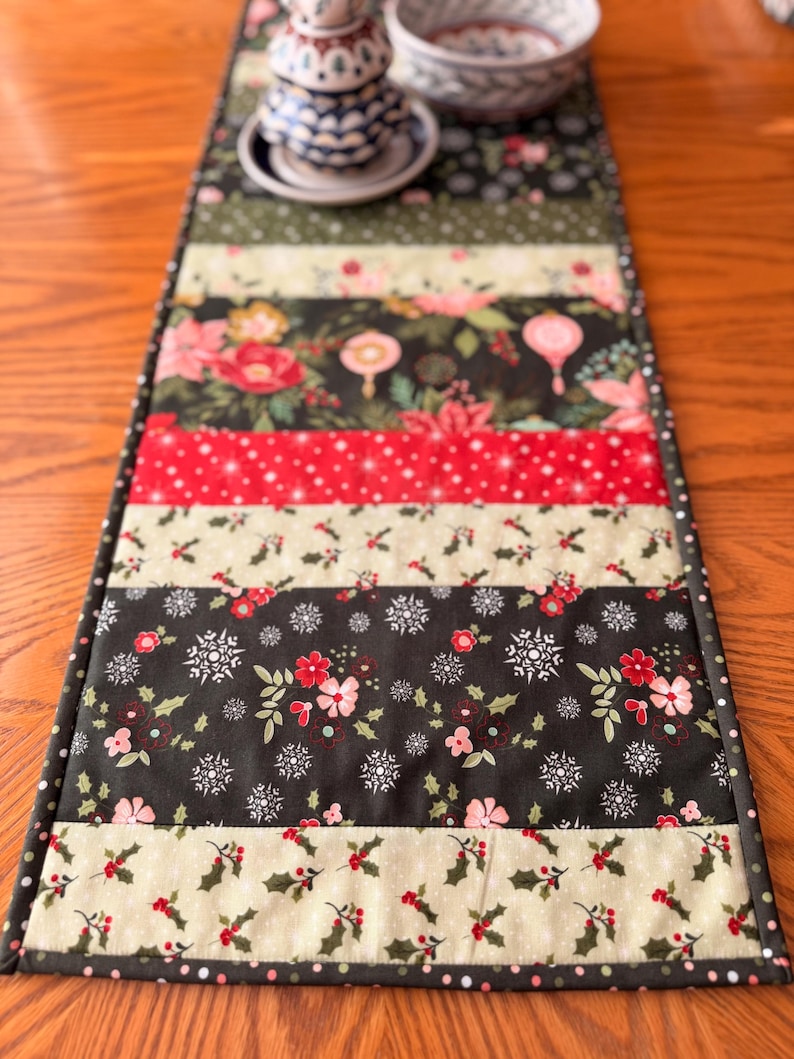 May include: A quilted table runner with a festive Christmas theme. The runner features alternating strips of fabric with floral, snowflake, and holly patterns in red, green, and cream colors. The edges are finished with a dark green polka dot border.