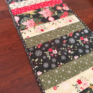 May include: A Christmas-themed table runner with a patchwork design. The runner features various fabrics with floral, snowflake, and dotted patterns in red, green, cream, and black. The runner is rectangular and appears to be handmade.