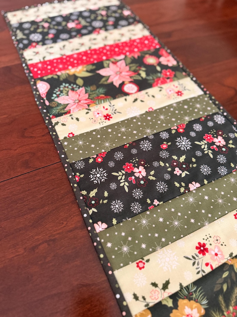 May include: A Christmas-themed table runner with a patchwork design. The runner features various fabrics with floral, snowflake, and dotted patterns in red, green, cream, and black. The runner is rectangular and appears to be handmade.