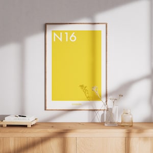 May include: A rectangular wall art print in a thin wooden frame featuring a solid bright yellow background. The text N16 is printed in white in the upper left corner, and the word LONDON is printed in small white letters at the bottom centre.