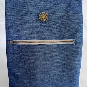 May include: A blue denim pouch with a zipper closure and a circular button. The button is gold-colored and has the words "Hello Sheng Jeans" printed on it. A black elastic loop is attached to the top.