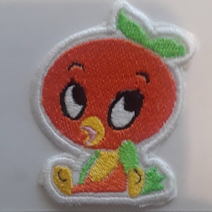 May include: Embroidered patch featuring a cartoon orange character with large eyes, a green leaf, and a white border. The character is orange with yellow and green accents. The patch is approximately 2 inches in height.
