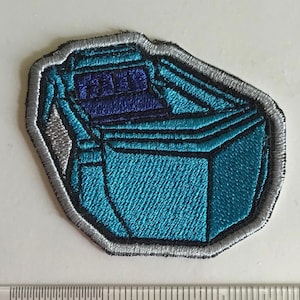 May include: Embroidered patch featuring a stylized, geometric design in shades of teal and dark blue, outlined in black and silver. The patch has a textured appearance, suggesting a fabric or textile base. The shape is an irregular polygon.