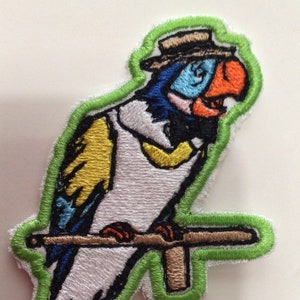 May include: Embroidered patch featuring a colorful parrot wearing a hat and perched on a branch. The parrot has blue, white, yellow, and orange details, outlined in green. The patch is designed with a white background and a green border.