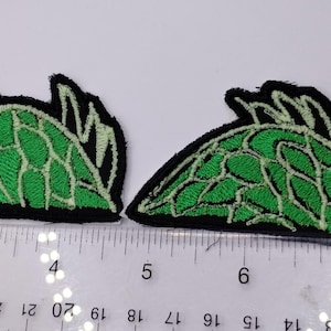 May include: Four embroidered patches depicting a green dragon in various poses. The patches are outlined in black and feature detailed stitching. A ruler is placed below the patches for scale.