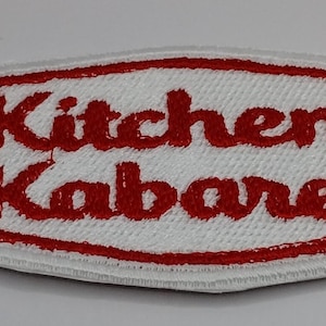May include: A white embroidered patch with a red border and the words "Kitchen Kabaret" in red script. The patch has a rectangular shape with rounded corners and a white background.