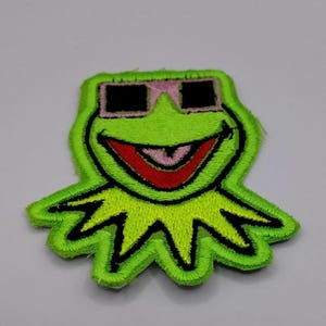 May include: Embroidered patch featuring a green frog with black sunglasses and a red mouth. The frog has a smiling expression and is outlined in black. The patch is a fun, colorful accessory.