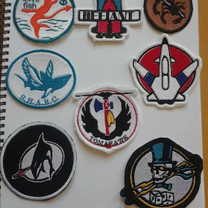 May include: A collection of embroidered patches with various designs. The patches feature images of a devil fish, a crab, a shark, a jet, a dolphin, a Native American tomahawk, and a skeleton in a top hat. Some patches have text, including "DEFANT" and "S.H.A.R.C."