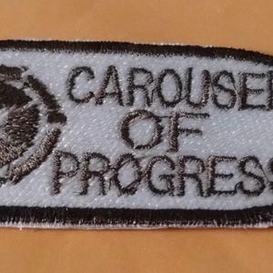 May include: An oval-shaped embroidered patch featuring a white background with a black border. On the left side is a circular gear-like icon in black and white. To the right, the text CAROUSEL OF PROGRESS is stitched in black capital letters.