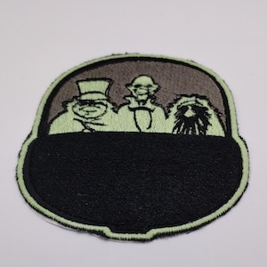 May include: Embroidered patch featuring three cartoon figures in a black and gray oval frame. The figures are in shades of green and white, with a black lower section. The patch has a green border and is likely for clothing or accessories.