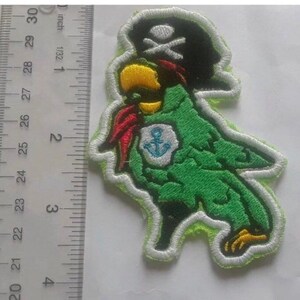 May include: Embroidered patch featuring a green parrot wearing a black pirate hat with a white skull and crossbones. The parrot has a yellow beak, red scarf, and a blue and white anchor emblem. The patch is approximately 3 inches in height.