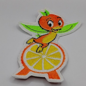 May include: Embroidered patch featuring a cartoon orange bird character with a smiling face, perched on a slice of orange. The patch has a white border, with orange, yellow, and green colors. The design is a whimsical, cheerful style.