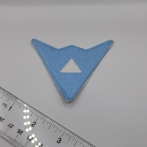 May include: A light blue embroidered patch with a white triangle in the center. The patch is in a stylized V-shape. A clear ruler is in the foreground.