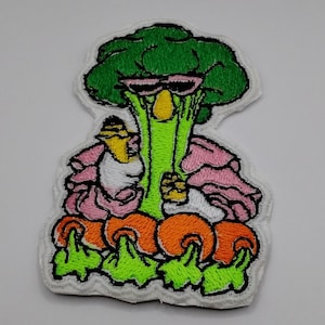 May include: An embroidered patch featuring a green broccoli character wearing black sunglasses. The character has a yellow nose, wears a pink ruffled garment, and holds a small yellow object. Below the character are four orange carrot shapes.