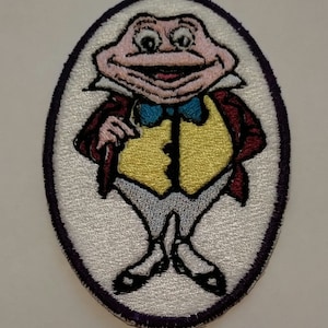 May include: An embroidered patch featuring a cartoon frog wearing a red jacket, yellow vest, and a blue bow tie. The frog has a large smile and is standing with its hands clasped in front of it.