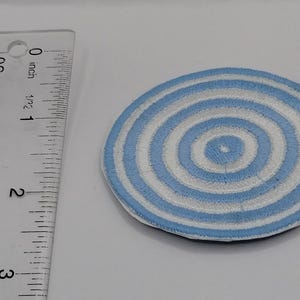 May include: A circular embroidered patch featuring concentric rings of light blue and white thread. A clear plastic ruler is positioned to the left, showing the patch has a diameter of approximately 2.5 inches across its widest point.