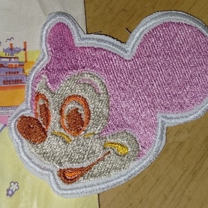 May include: A pink and white embroidered patch of a cartoon mouse with large ears. The mouse has a white face with orange eyes and a yellow smile.