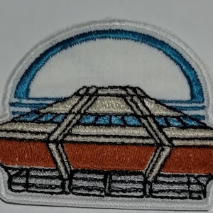 May include: Embroidered patch featuring a stylized architectural design with an orange base, a white and blue upper section, and a blue arched top. The patch has a white border and black stitching, suitable for jackets, bags, or other fabric items.
