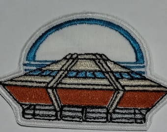 May include: Embroidered patch featuring a stylized architectural design with an orange base, a white and blue upper section, and a blue arched top. The patch has a white border and black stitching, suitable for jackets, bags, or other fabric items.