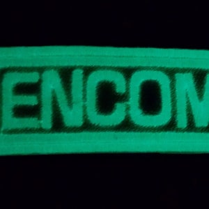 May include: Glow-in-the-dark rectangular patch with the word "ENCOM" in bold, green letters. The patch has a raised border and is set against a black background. The patch is likely for clothing or gear.