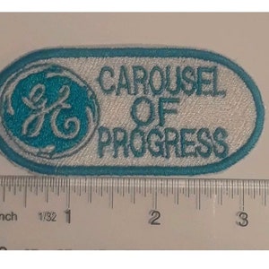 May include: Embroidered patch with a teal and white design. The patch features the text "CAROUSEL OF PROGRESS" in white, with a teal logo on the left side. The patch is oval-shaped with a teal border.