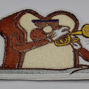 May include: Embroidered patch featuring a slice of bread playing a trumpet. The bread slice is brown with a cream-colored center, wearing a purple visor and white gloves. The trumpet is gold, and the patch has a white border.