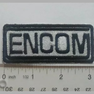 May include: A black and white embroidered patch with the word "ENCOM" in white lettering on a black background. The patch is approximately 1 inch wide.
