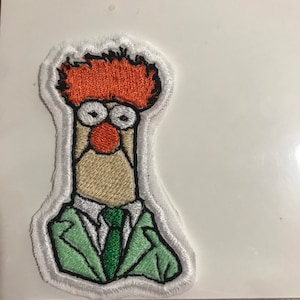 May include: Embroidered patch featuring a cartoon character with orange hair, a red nose, and large, round eyes. The character wears a green suit and tie. The patch is outlined in white and measures approximately 2 inches in height.