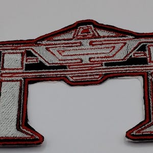 May include: An embroidered patch featuring a geometric, arch-like design in silver and black with a red border. A clear plastic ruler is positioned to the left, showing the patch width is approximately 3.5 inches and height is 2.5 inches.