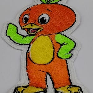 May include: Embroidered patch featuring a cartoon orange character with green arms and a yellow belly. The character has a smiling face and is waving. The patch has a white border.
