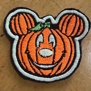 May include: An embroidered patch shaped like a pumpkin with mouse-like ears. The pumpkin is orange with black outlines, a green leaf on top, and a light blue border. The face features light blue eyes, a light blue nose, and a curved mouth.