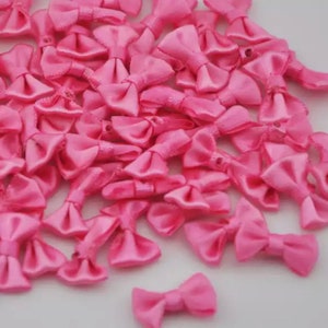May include: A collection of small, bright pink satin bows. The bows are neatly tied and have a shiny, smooth texture. They are scattered across a white surface, creating a visually appealing arrangement. These bows are ideal for crafting and embellishments.