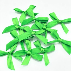 May include: A collection of small, bright green satin bows. Each bow is tied in the center, with four loops extending outwards. The bows are scattered on a white surface, creating a cheerful and decorative display.