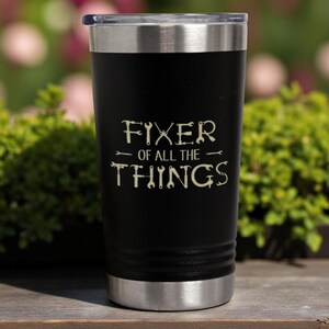 May include: Black insulated tumbler with a silver rim and lid. The tumbler has the text "FIXER OF ALL THE THINGS" in a light beige color, with tool graphics. The tumbler is sitting on a wooden surface with a green and pink background.