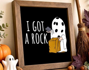 May include: A square wooden frame containing a black sign with white text that reads I GOT A ROCK. The graphic features a white ghost character with black spots holding a gray rock and a brown paper bag, depicted in a simple cartoon style.